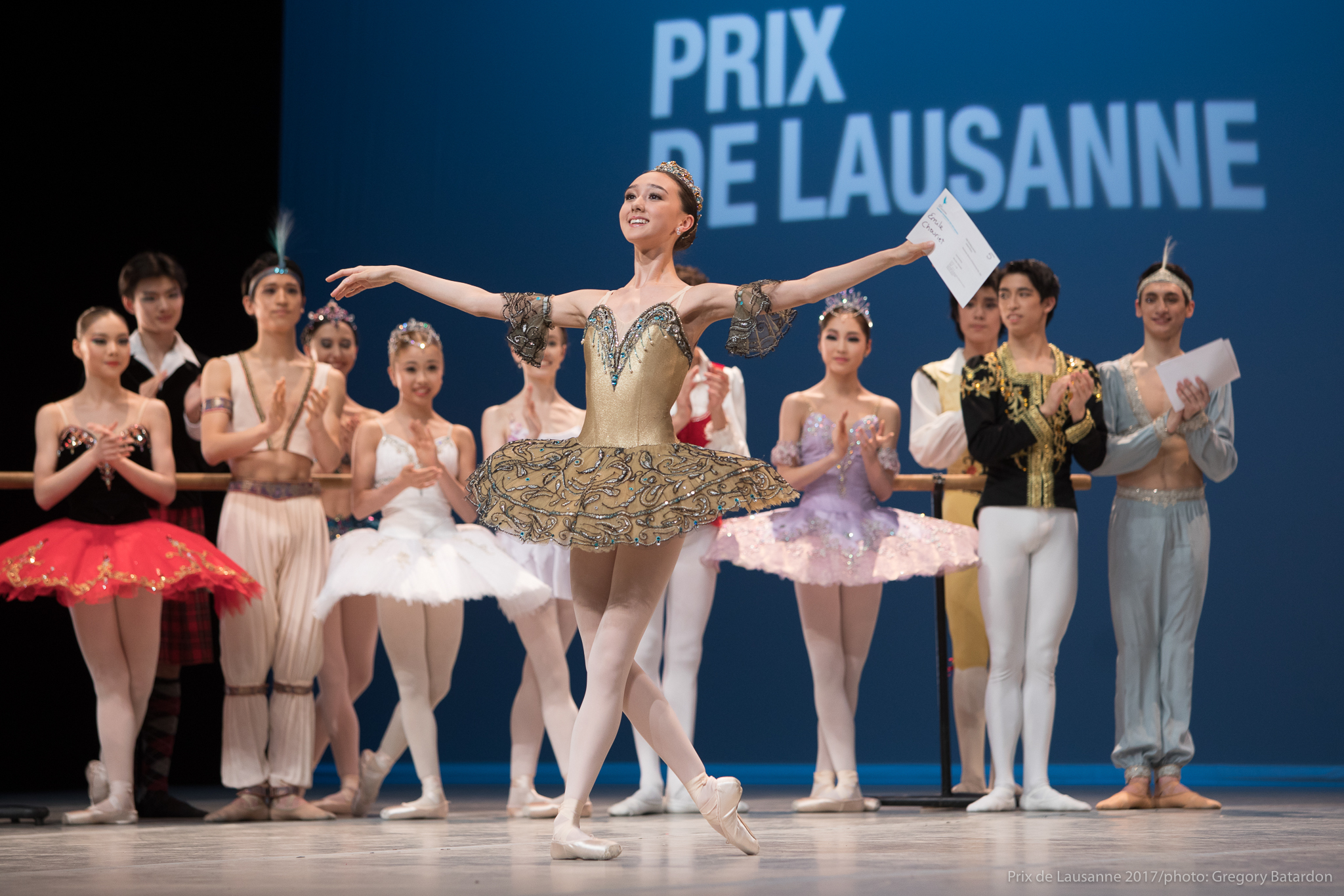 Danseuse en tutu sur scène exéctant une pose gracieuse lors du Prix de Lausanne, entourée d'autres participants en tenue de ballet.