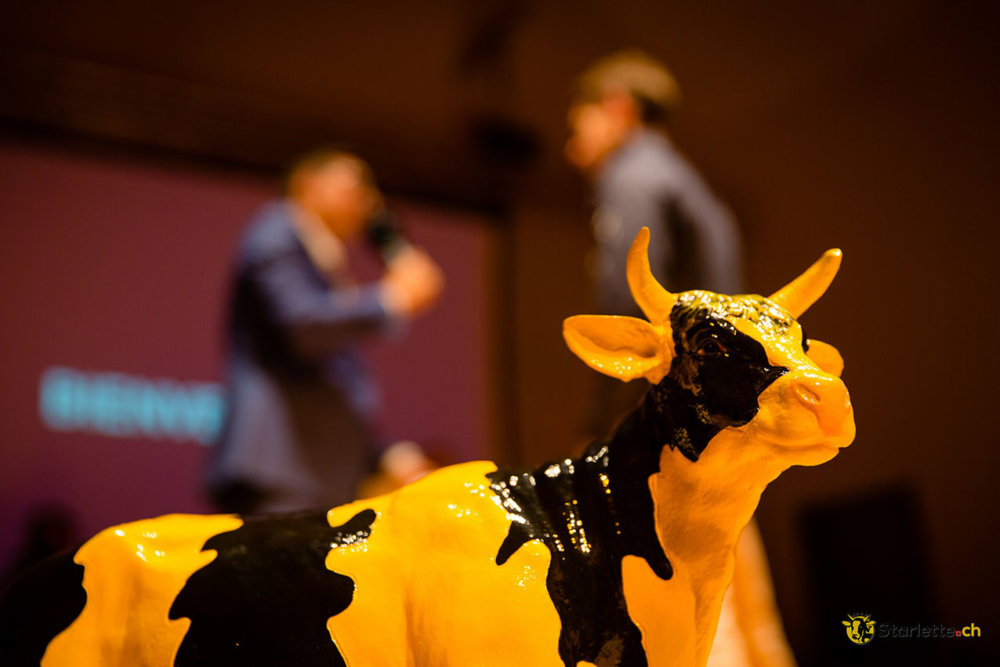 Figurine de vache jaune et noire mise en avant sur une scène, avec deux personnes floues en arrière-plan lors d'un événement ou d'une présentation.
