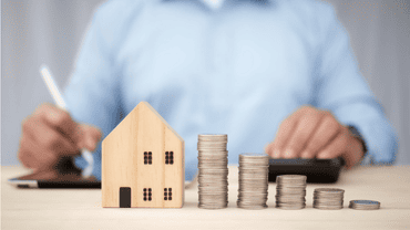 Une personne en chemise bleue, floue à l'arrière-plan, utilise une calculatrice devant une petite maison en bois et des pièces de monnaie alignées sur une table, symbolisant l'investissement immobilier et la gestion financières.