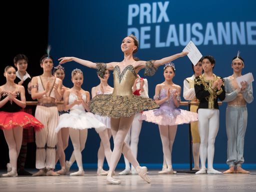 Danseuse en tutu sur scène exéctant une pose gracieuse lors du Prix de Lausanne, entourée d'autres participants en tenue de ballet.
